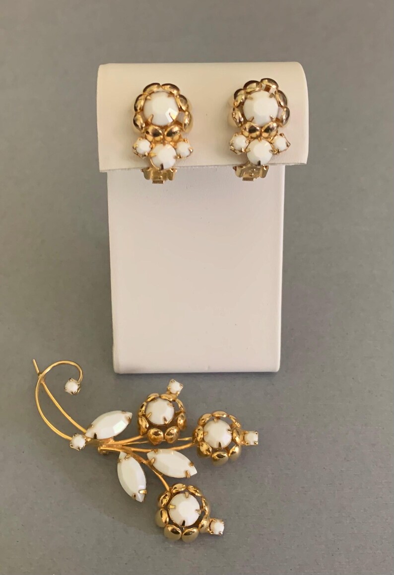 May include: A set of vintage jewelry featuring a pair of clip-on earrings and a brooch. The earrings and brooch have a gold-tone metal setting with white, round, and oval-shaped accents. The earrings are mounted on a white display stand.
