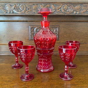 May include: A vintage red glass decanter set with a stopper and four matching wine glasses. The decanter and glasses have a textured pattern. The set is displayed on a wooden surface.