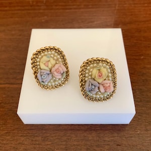 May include: A pair of vintage clip-on earrings. Each earring features a gold-tone rope-like border surrounding a cluster of small, pastel-colored porcelain roses. The roses are set on a bed of tiny faux pearls.