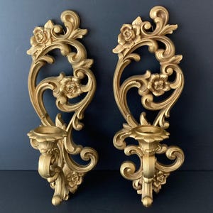 May include: A pair of gold-colored wall sconces featuring intricate scrollwork and floral motifs. Each sconce has a central candle holder cup integrated into the ornate, elongated frame, showcasing detailed relief carvings of leaves and blossoms.
