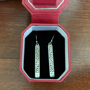Puede incluir: Pendientes colgantes plateados con dise&ntilde;o rectangular. Cada pendiente presenta un intrincado patr&oacute;n de volutas y una peque&ntilde;a gema transparente en la parte inferior. Los pendientes se exhiben en una caja de joyer&iacute;a roja con interior negro.