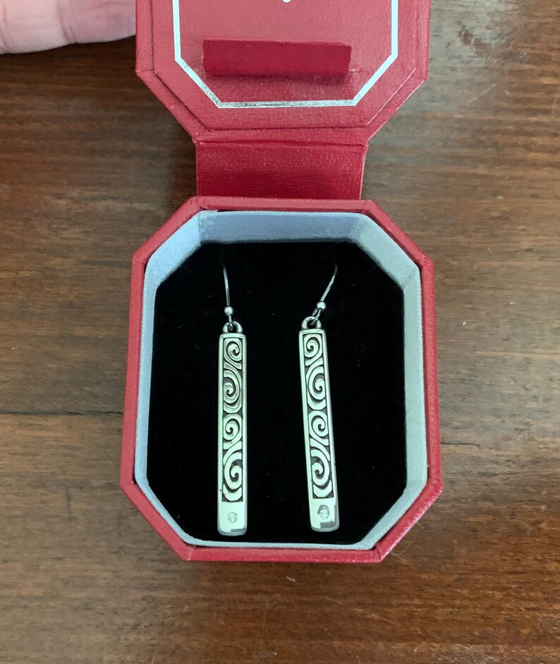 Puede incluir: Pendientes colgantes plateados con dise&ntilde;o rectangular. Cada pendiente presenta un intrincado patr&oacute;n de volutas y una peque&ntilde;a gema transparente en la parte inferior. Los pendientes se exhiben en una caja de joyer&iacute;a roja con interior negro.