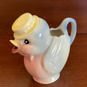 May include: A blue and pink ceramic creamer in the shape of a bird wearing a yellow hat. The bird has a black eye and a pink beak.