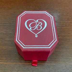 Puede incluir: Una caja de joyer&iacute;a roja, octogonal, con un borde blanco y un logotipo en forma de coraz&oacute;n con la letra "B". La caja tiene un peque&ntilde;o tirador de cinta roja. La caja est&aacute; cerrada y se encuentra sobre una superficie de madera.