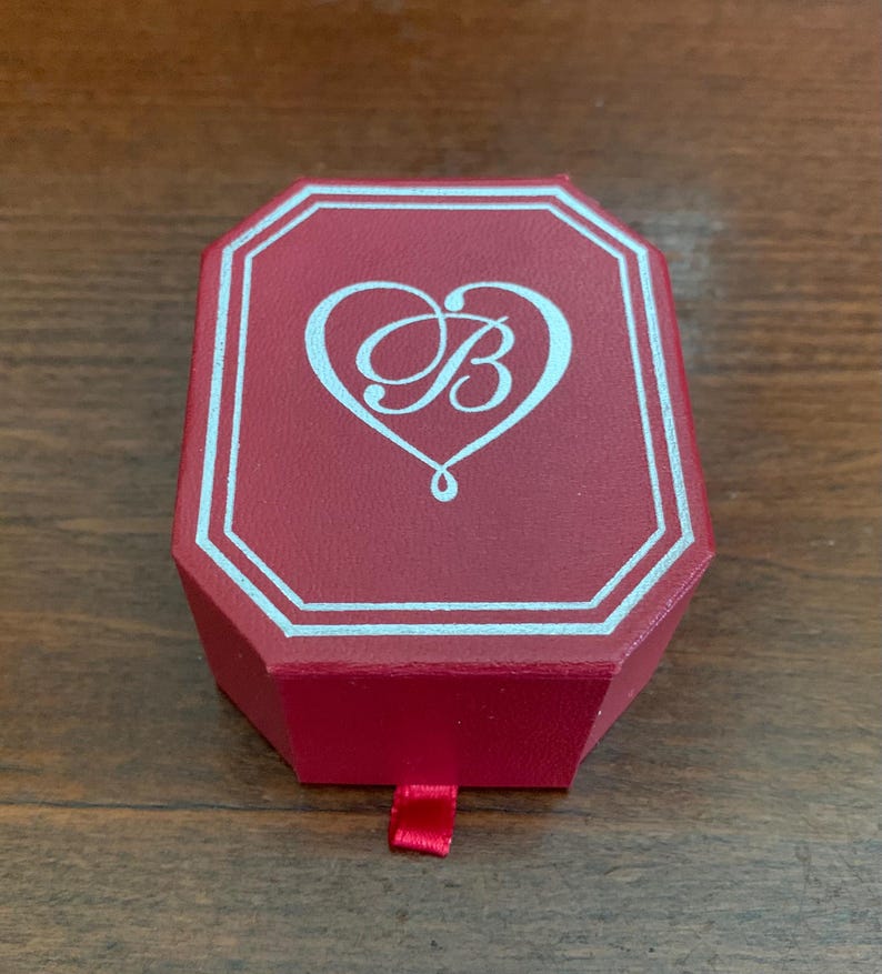 Puede incluir: Una caja de joyer&iacute;a roja, octogonal, con un borde blanco y un logotipo en forma de coraz&oacute;n con la letra "B". La caja tiene un peque&ntilde;o tirador de cinta roja. La caja est&aacute; cerrada y se encuentra sobre una superficie de madera.