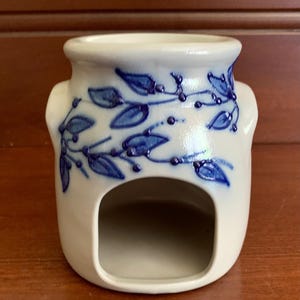 May include: A white ceramic oil warmer featuring a hand-painted blue vine and leaf pattern. The cylindrical vessel has a wide opening at the top and a large, arched cutout at the base to accommodate a heat source, with a smooth, glossy finish.