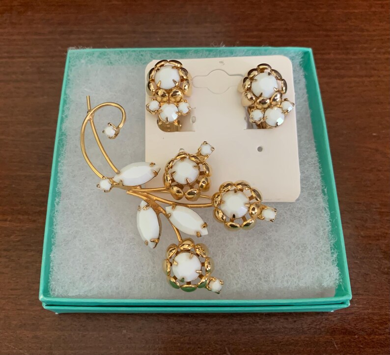 May include: A vintage jewelry set featuring a gold-tone brooch and matching clip-on earrings. The brooch has a floral design with white cabochon stones and gold accents. The earrings also have a floral design with white stones.