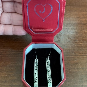 Puede incluir: Un par de pendientes plateados con dise&ntilde;o rectangular y patr&oacute;n de espirales, presentados en un joyero rojo abierto. La caja tiene un dise&ntilde;o de coraz&oacute;n en la tapa. Los pendientes est&aacute;n sobre un fondo de terciopelo negro.