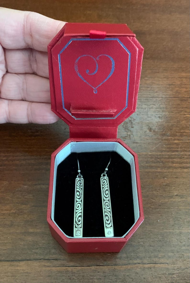 Puede incluir: Un par de pendientes plateados con dise&ntilde;o rectangular y patr&oacute;n de espirales, presentados en un joyero rojo abierto. La caja tiene un dise&ntilde;o de coraz&oacute;n en la tapa. Los pendientes est&aacute;n sobre un fondo de terciopelo negro.