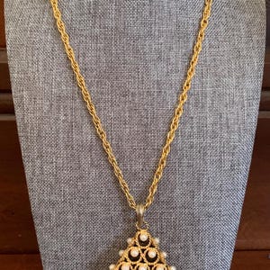 May include: A gold chain necklace with a triangular pendant featuring a geometric design of gold wire and white pearls.