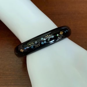 May include: A black bangle bracelet with inlaid floral and geometric designs. The bracelet features gold, blue, and iridescent accents. The bracelet is shown on a white surface.