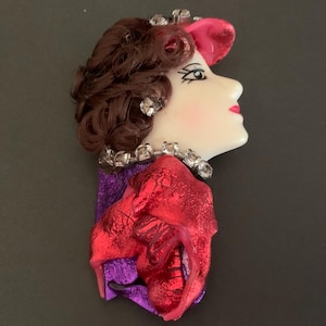 May include: A brooch featuring a profile of a face with brown hair, a red hat, and a red garment. The figure wears a clear rhinestone necklace and earring. The face is cream-colored with painted features, including dark eyelashes and red lips.