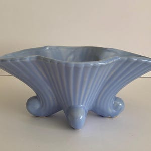 May include: A light blue ceramic vase with a shell-like design. The vase has a square shape with curved edges and three rounded feet. The exterior features vertical ridges, adding texture to the piece. A decorative item.