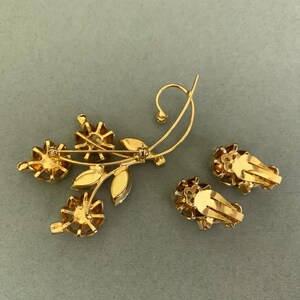 May include: A gold-toned floral brooch and matching clip-on earrings. The brooch features three flower-shaped accents with amber-colored stones, along with leaves and a curved stem. The earrings have a similar flower design.
