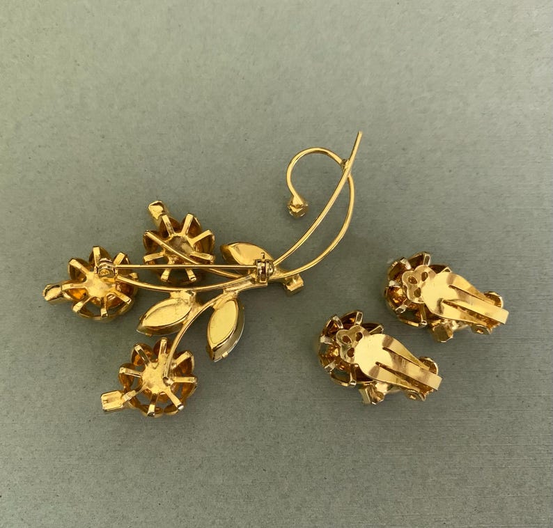 May include: A gold-toned floral brooch and matching clip-on earrings. The brooch features three flower-shaped accents with amber-colored stones, along with leaves and a curved stem. The earrings have a similar flower design.