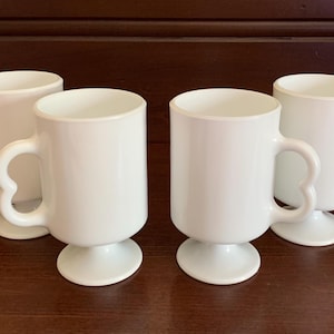 May include: A set of four white milk glass mugs, each featuring a smooth cylindrical body, a pedestal base, and a distinctive heart-shaped handle. These opaque white drinking vessels are arranged in a group, showcasing their uniform design.