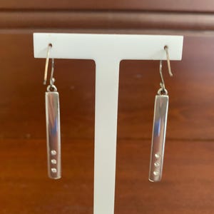 Puede incluir: Un par de pendientes colgantes plateados. Cada pendiente presenta una barra rectangular con tres peque&ntilde;as piedras redondas y transparentes. Los pendientes cuelgan de un gancho sencillo. Los pendientes se exhiben en un soporte blanco para pendientes.