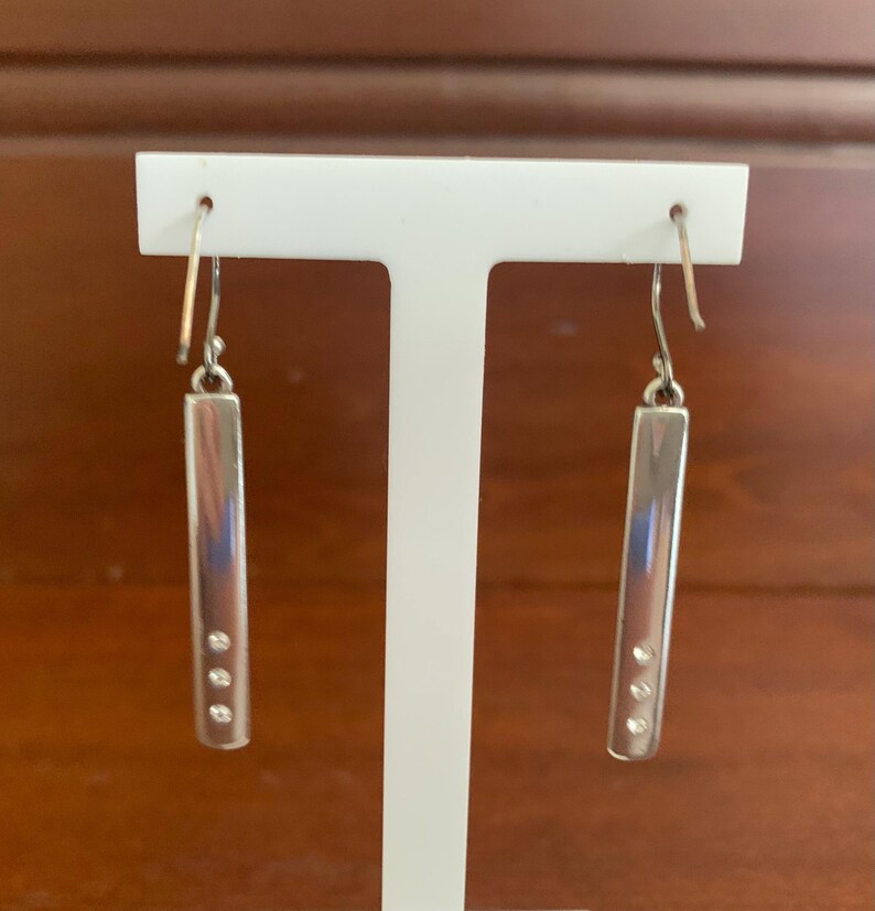 Puede incluir: Un par de pendientes colgantes plateados. Cada pendiente presenta una barra rectangular con tres peque&ntilde;as piedras redondas y transparentes. Los pendientes cuelgan de un gancho sencillo. Los pendientes se exhiben en un soporte blanco para pendientes.