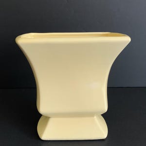 May include: A pale yellow ceramic vase with a square top and a flared body. The vase has a pedestal base and a smooth, glossy finish. The vase is a decorative item.