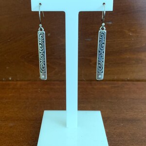 Puede incluir: Pendientes colgantes plateados exhibidos en un soporte blanco. Cada pendiente presenta un dise&ntilde;o rectangular con un intrincado patr&oacute;n de remolino y una peque&ntilde;a gema transparente. Los pendientes cuelgan de un gancho simple.