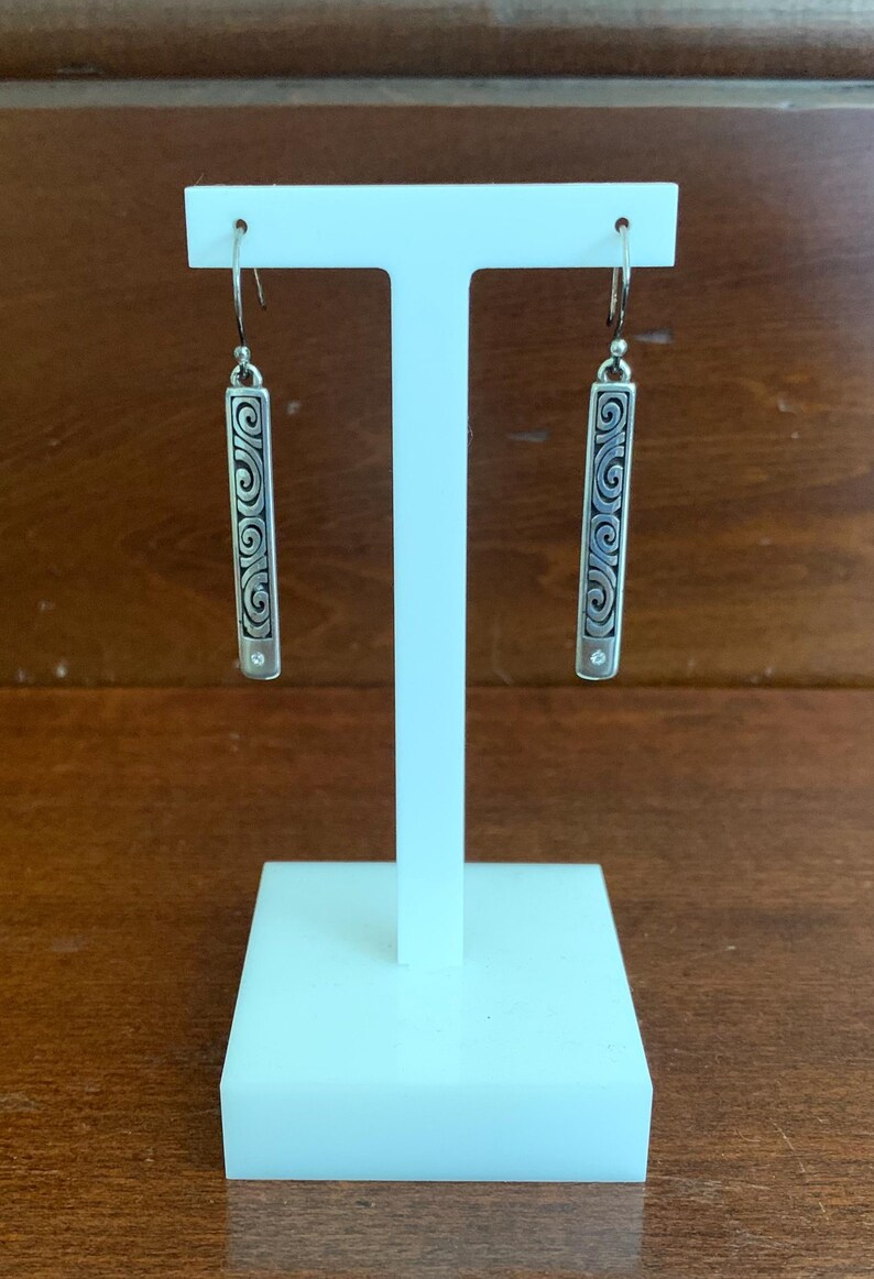 Puede incluir: Pendientes colgantes plateados exhibidos en un soporte blanco. Cada pendiente presenta un dise&ntilde;o rectangular con un intrincado patr&oacute;n de remolino y una peque&ntilde;a gema transparente. Los pendientes cuelgan de un gancho simple.