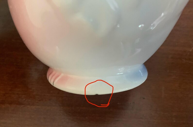 May include: A white ceramic vase with a pink and blue ombre glaze. The vase has a round body and a flared lip. The base of the vase has a small chip.