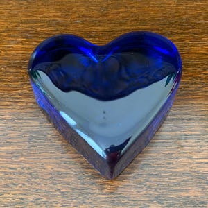 May include: A heart-shaped, cobalt blue glass paperweight. The glass is smooth and reflective, with a deep, rich color. The heart is sitting on a wooden surface.