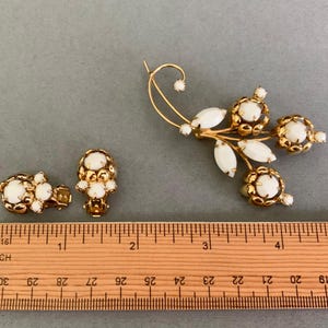 May include: Vintage gold-tone jewelry set featuring a floral brooch and matching clip-on earrings. The brooch has a curved design with white oval and round stones. The earrings have a cluster of white stones. The ruler shows the brooch is approximately 4 inches long.
