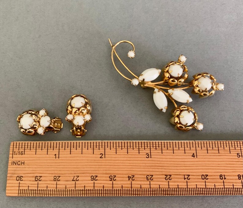 May include: Vintage gold-tone jewelry set featuring a floral brooch and matching clip-on earrings. The brooch has a curved design with white oval and round stones. The earrings have a cluster of white stones. The ruler shows the brooch is approximately 4 inches long.