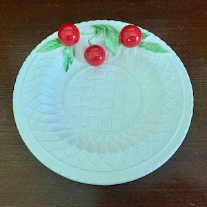 May include: A white ceramic plate with a raised cherry design. The plate features three red cherries with green leaves and a woven pattern in the center. The rim has a decorative edge.