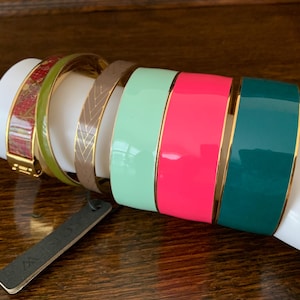 Op de afbeelding: Een verzameling kleurrijke armbanden tentoongesteld op een witte mannequinarm. De armbanden hebben verschillende ontwerpen en kleuren, waaronder rood, groen, bruin, mintgroen, felroze en blauwgroen. Elke armband heeft een goudkleurige rand. Een zwart label is zichtbaar.