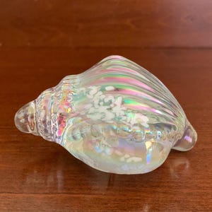 May include: A clear glass seashell figurine featuring an iridescent finish that reflects rainbow colors. The shell has a spiral shape with raised ridges and internal white speckled inclusions throughout the body, creating a textured appearance.