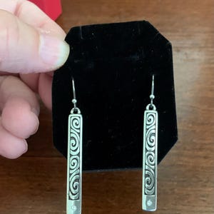 Puede incluir: Pendientes colgantes plateados. Cada pendiente presenta una barra rectangular con un dise&ntilde;o de espirales decorativas y una peque&ntilde;a gema transparente en la parte inferior. Los pendientes se exhiben sobre una superficie de terciopelo negro.
