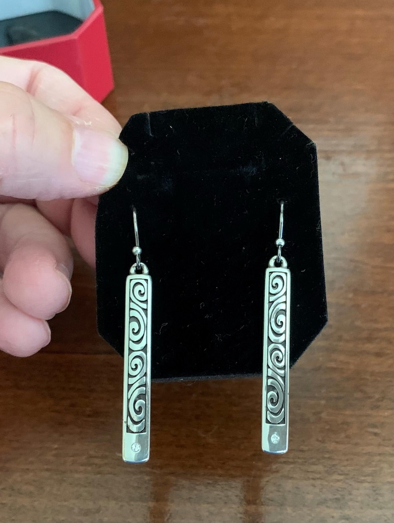 Puede incluir: Pendientes colgantes plateados. Cada pendiente presenta una barra rectangular con un dise&ntilde;o de espirales decorativas y una peque&ntilde;a gema transparente en la parte inferior. Los pendientes se exhiben sobre una superficie de terciopelo negro.