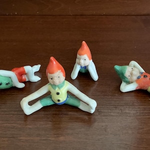 May include: Four small ceramic elf figurines in various poses. One lies on its side with a yellow hat, one sits upright with an orange hat, one is in a split position with an orange hat, and one lies on its back with a green hat and blue shorts.