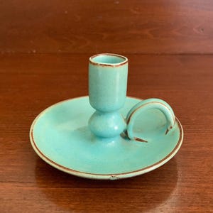 May include: A turquoise ceramic candle holder with a circular base, a central cylindrical socket for a taper candle, and a small curved handle on the side. The piece features a smooth, matte finish with subtle brown edges along the rim and base.