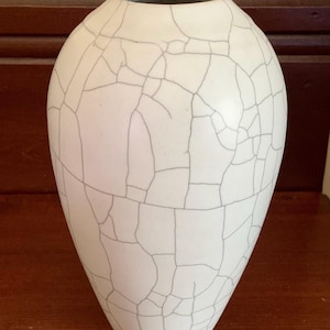 May include: A white ceramic vase with a tapered, rounded silhouette and a narrow opening at the top. The exterior surface features a distinct dark gray crackle glaze pattern that creates a network of irregular lines across the entire vessel.