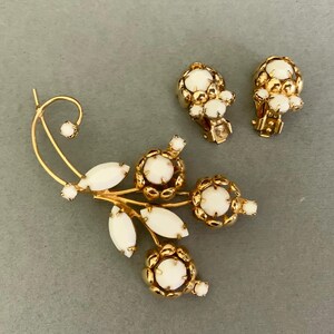 May include: A vintage jewelry set featuring a gold-toned brooch and matching clip-on earrings. The brooch has a floral design with white oval and round accents. The earrings and brooch have a gold filigree design around the white accents.