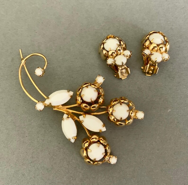May include: A vintage jewelry set featuring a gold-toned brooch and matching clip-on earrings. The brooch has a floral design with white oval and round accents. The earrings and brooch have a gold filigree design around the white accents.