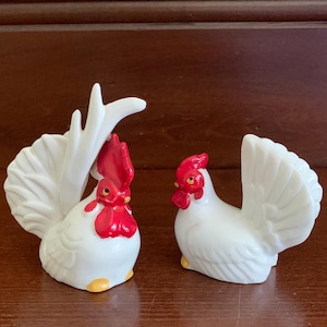 May include: A pair of ceramic rooster figurines. The roosters are white with red combs and wattles, and yellow feet. One rooster has a tall, stylized tail. The other rooster has a fanned tail.
