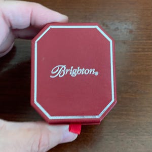 Puede incluir: Una caja octogonal roja con un borde plateado y la palabra "Brighton" en escritura cursiva. La caja tiene un peque&ntilde;o tirador de cinta roja. La caja est&aacute; sujeta por una mano.