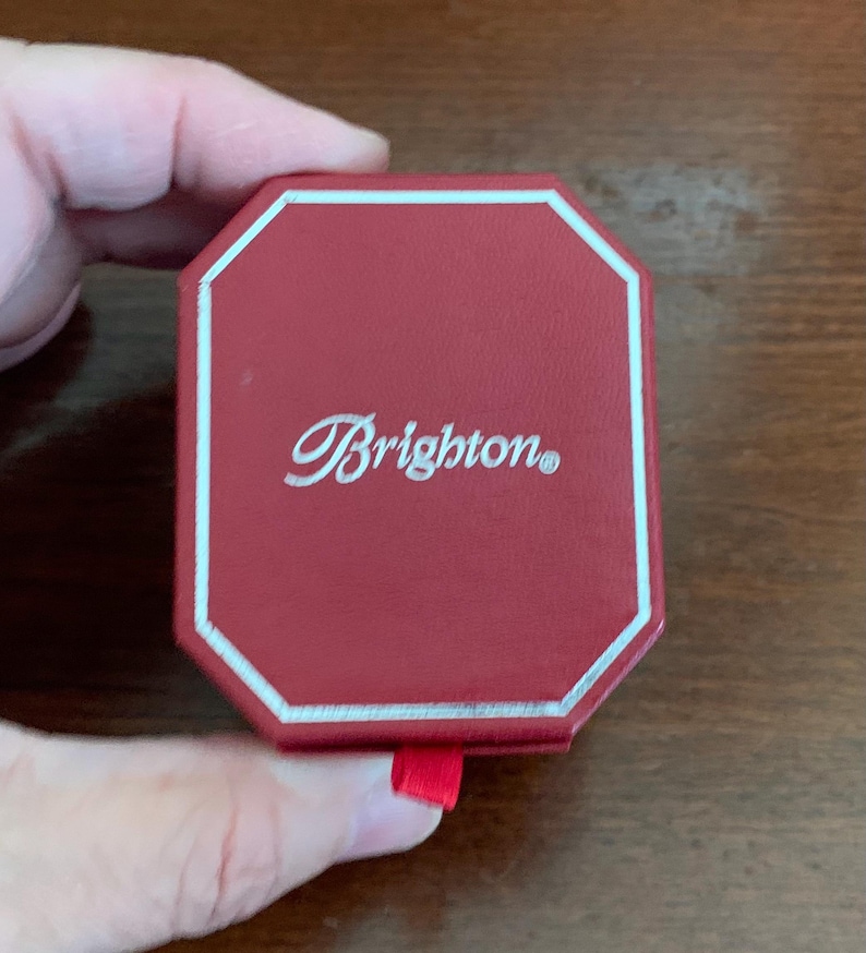 Puede incluir: Una caja octogonal roja con un borde plateado y la palabra "Brighton" en escritura cursiva. La caja tiene un peque&ntilde;o tirador de cinta roja. La caja est&aacute; sujeta por una mano.