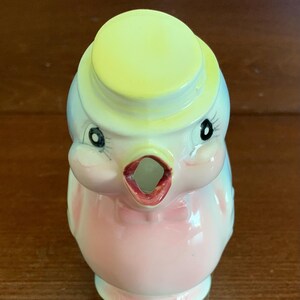 May include: A pink and blue ceramic bird pitcher with a yellow hat. The bird has a black bow tie and black eyes. The pitcher has a spout shaped like a bird's beak.