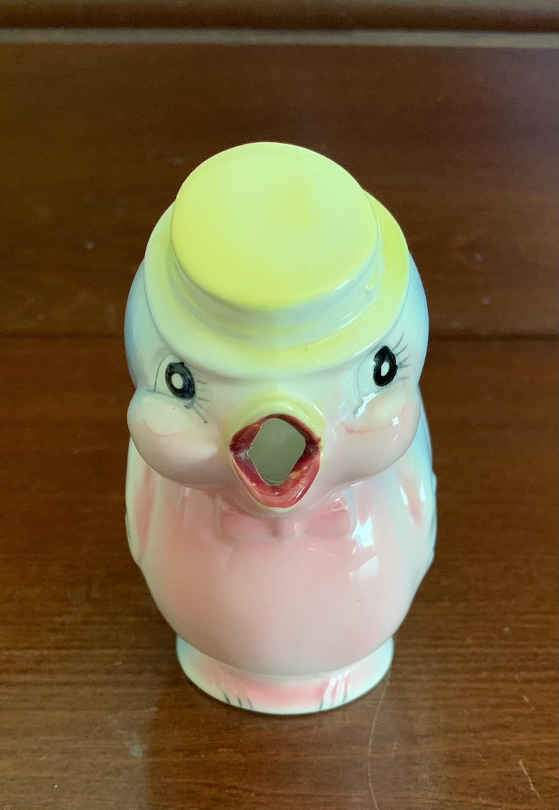 May include: A pink and blue ceramic bird pitcher with a yellow hat. The bird has a black bow tie and black eyes. The pitcher has a spout shaped like a bird's beak.