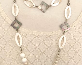 May include: A layered necklace featuring white oval and diamond-shaped shell pendants, interspersed with small beads and connected by metal links. The necklace is displayed on a white lace background, showcasing its delicate design.