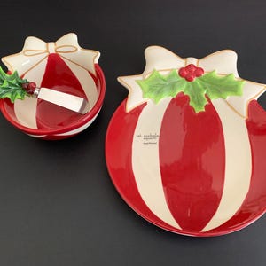 May include: A festive Christmas serving set featuring a bowl with a spreader and a plate. Both pieces are red and white striped, shaped like ornaments, and adorned with holly and bow details. The set is perfect for holiday entertaining.