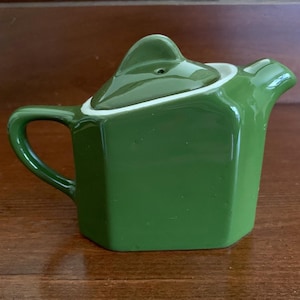May include: A small, glossy, olive green ceramic teapot with a hexagonal body, a curved handle, and a matching lid featuring a triangular finial. The rim of the teapot opening is white, providing a clean contrast against the solid green glaze.