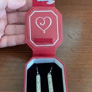 Puede incluir: Pendientes plateados con dise&ntilde;o rectangular y patr&oacute;n de espiral, presentados en una caja octogonal roja. La caja tiene un dise&ntilde;o de coraz&oacute;n en la tapa y un interior de terciopelo negro. Los pendientes est&aacute;n sobre una superficie de madera.