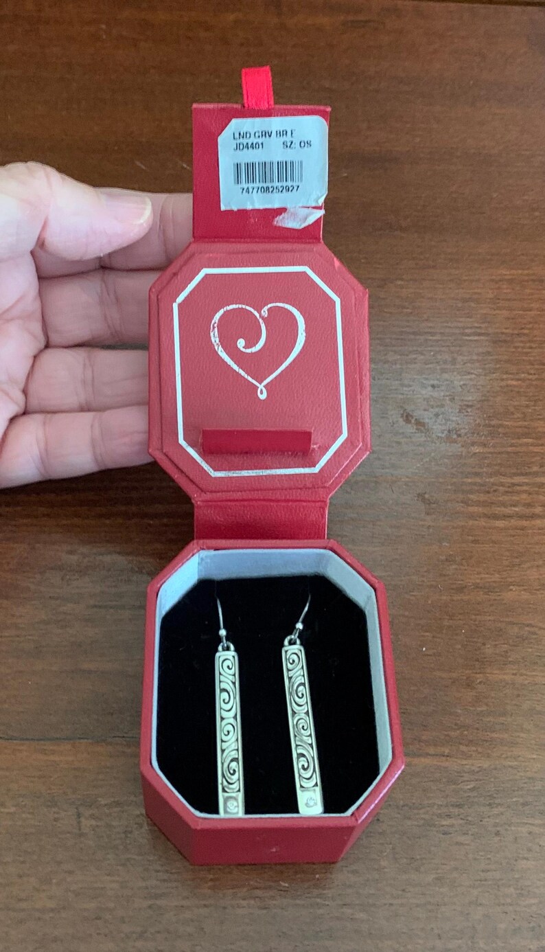 Puede incluir: Pendientes plateados con dise&ntilde;o rectangular y patr&oacute;n de espiral, presentados en una caja octogonal roja. La caja tiene un dise&ntilde;o de coraz&oacute;n en la tapa y un interior de terciopelo negro. Los pendientes est&aacute;n sobre una superficie de madera.