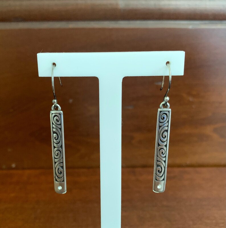 Puede incluir: Pendientes colgantes plateados con dise&ntilde;o rectangular. Cada pendiente presenta un patr&oacute;n de remolino grabado y una peque&ntilde;a piedra transparente en la parte inferior. Los pendientes se exhiben sobre un soporte blanco sobre un fondo marr&oacute;n.